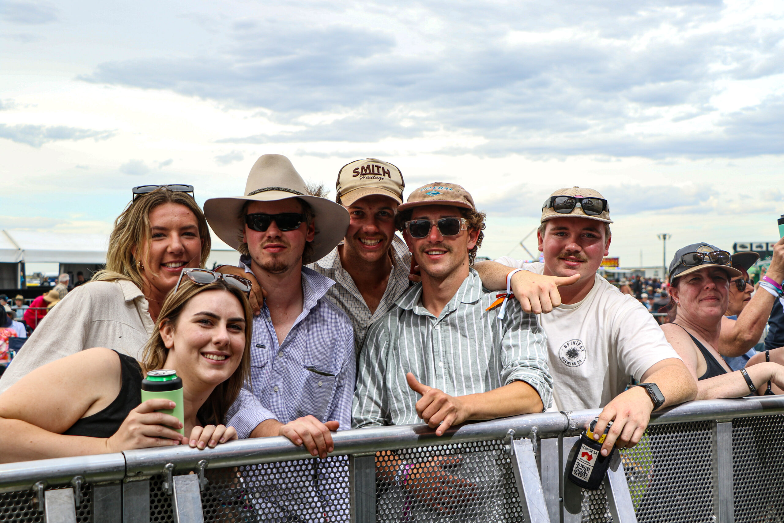 Group of six smiling individuals wearing hats and casual attire at an outdoor event, some holding drinks, with crowds and tents in the background at The Bend Classic.