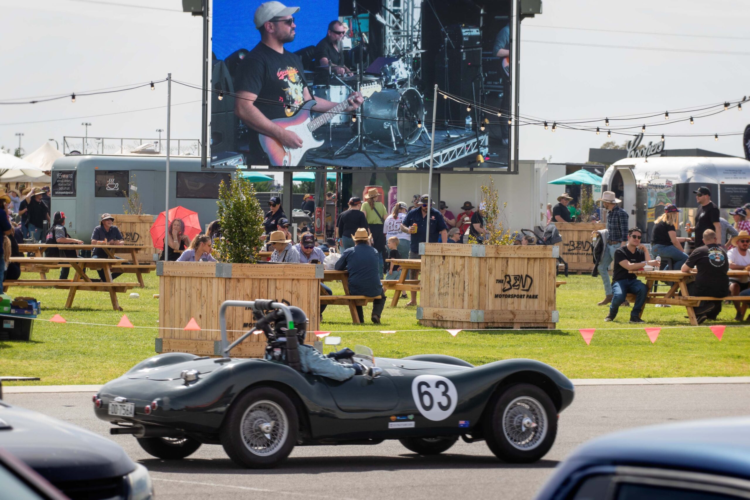 A vintage sports car with the number 63 drives past an outdoor event where people are seated at wooden benches. A large screen displays a live band performance at The Bend Classic.